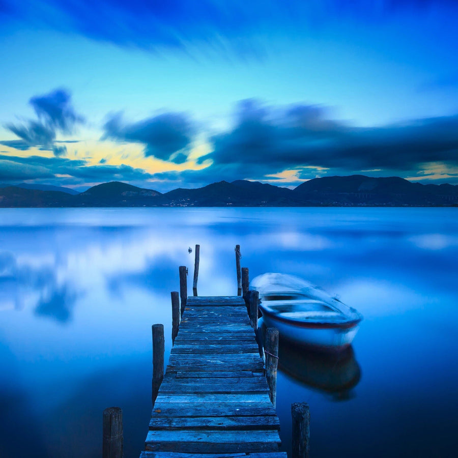 A serene lakeside view at twilight features a wooden dock extending into calm blue waters and a small rowboat, reminiscent of the Jetty and Boat Wall Mural by Deposit Photo. The sky has wispy clouds and distant hills in hues of blue and soft yellow, creating a tranquil scene.