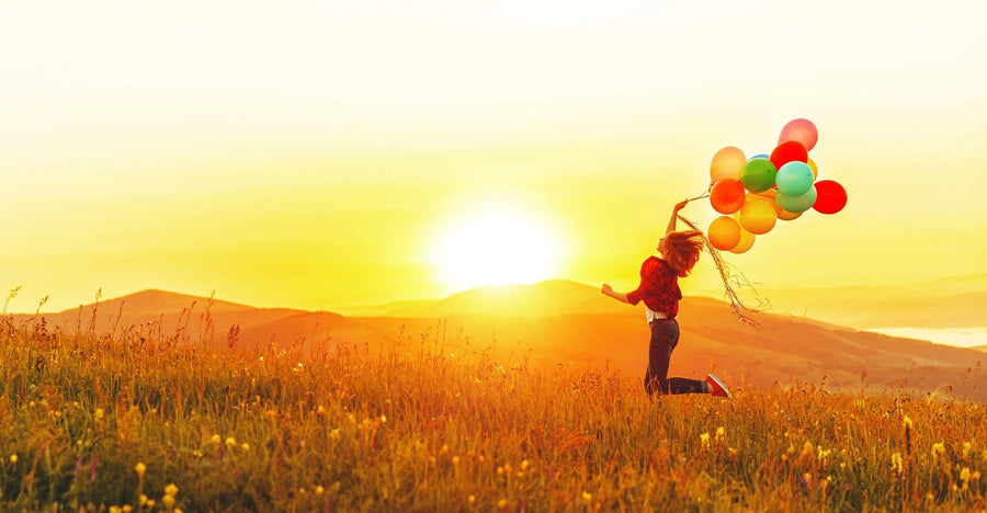 Running joyfully through a grassy field at sunset, a person clutches colorful balloons, resembling the vibrant serenity of the Joyful Sunset Freedom Wall Mural with its warm glow and nature-inspired scenery of gentle hills and wildflowers.