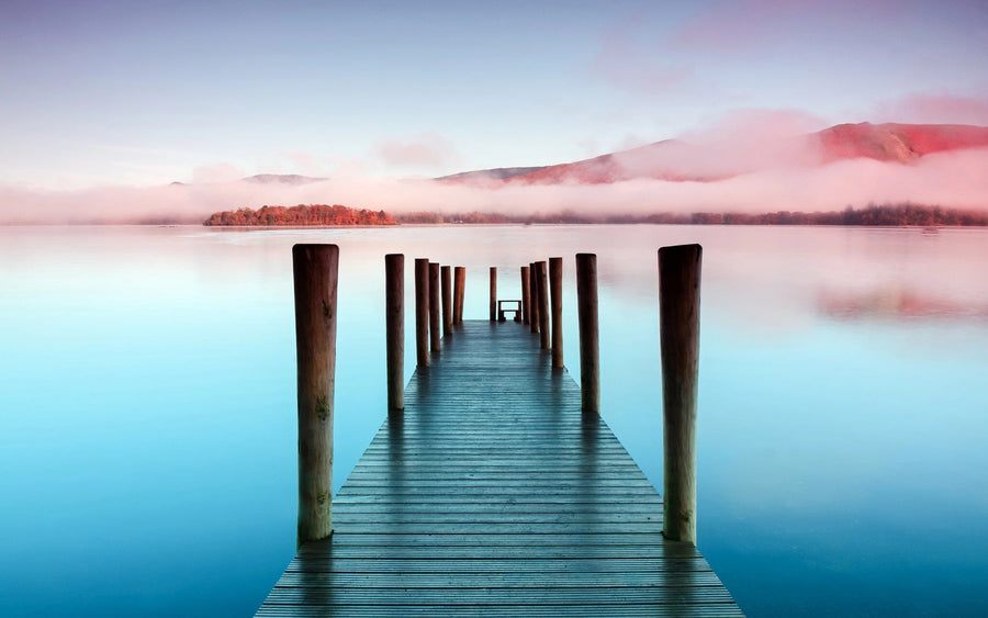 A wooden dock extends into a calm lake under misty skies as softly glowing hills reflect on the water, creating a tranquil scene. The Misty Lake Morning Wall Mural is ideal for custom prints or as a peel and stick wall mural.