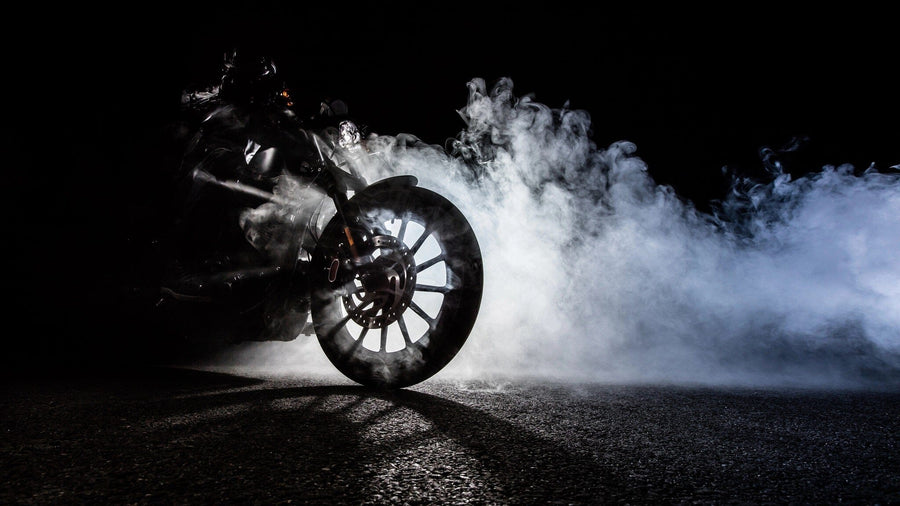 The Motorcycle Burnout Smoke Wall Mural from Deposit Photo depicts a dramatic scene of a motorcycle performing a burnout on a dark surface, emitting thick white smoke. The prominent front wheel and reflective rim create striking contrast against the black background.