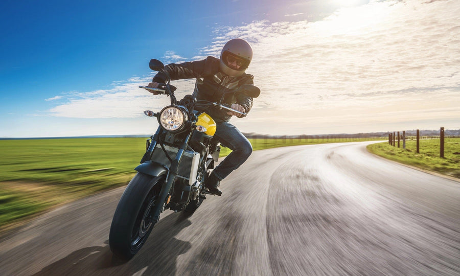 A motorcyclist in a black helmet and leather jacket leans into a curve on a rural road, embodying the thrilling allure of the Motorcycle Rider Adventure Wall Mural by Deposit Photo. The sun casts shadows as green fields and blue sky mirror the mural's sense of speed and freedom.