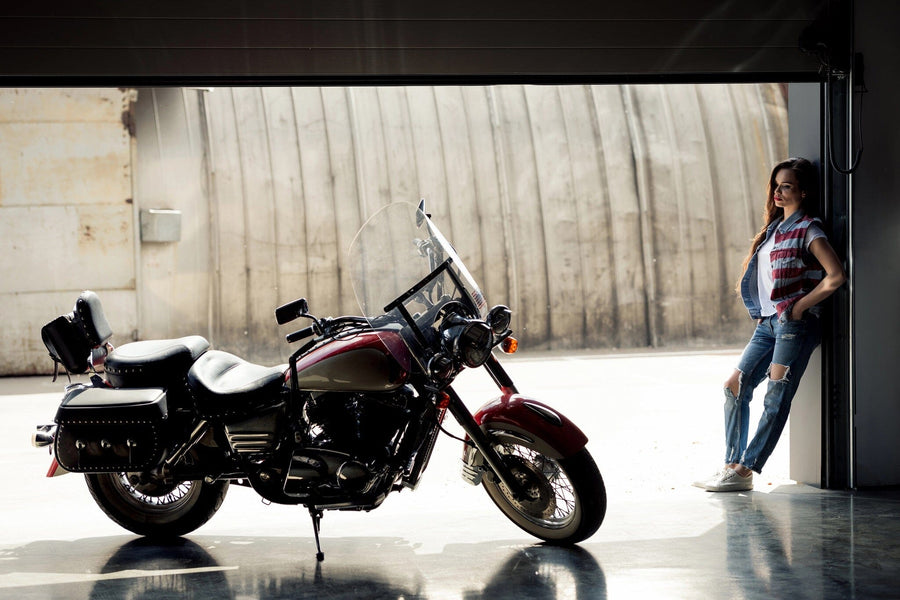 A woman in plaid and ripped jeans leans against a garage door, admiring a red motorcycle inside. The sunlight highlights the bike's sleek design, evoking the "Motorcycle and Rider Wall Mural" by Deposit Photo, known for its artful details in living spaces.