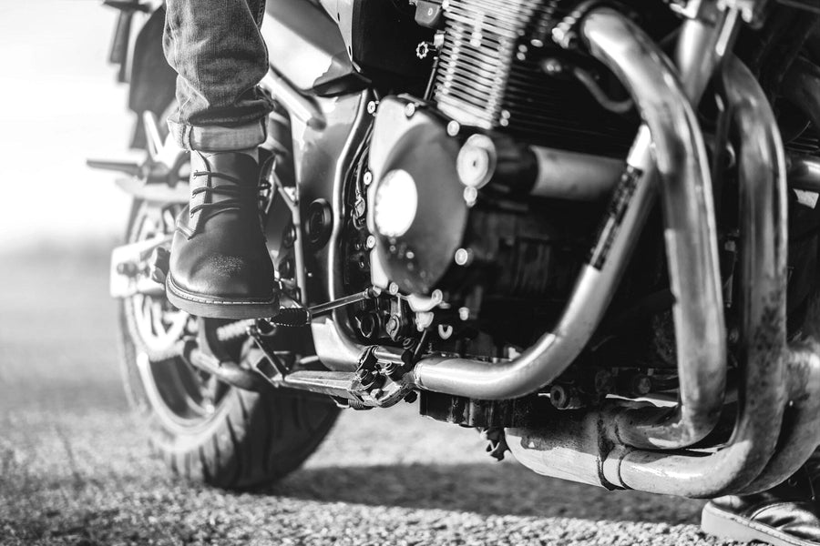 The "Motorcycle Rider Boot Wall Mural" by Deposit Photo captures the adventurous spirit with a black-and-white close-up of a motorcycle rider's boot on the footrest. The detailed engine and exhaust pipes cast shadows, and the blurred road background suggests motion or readiness to ride.