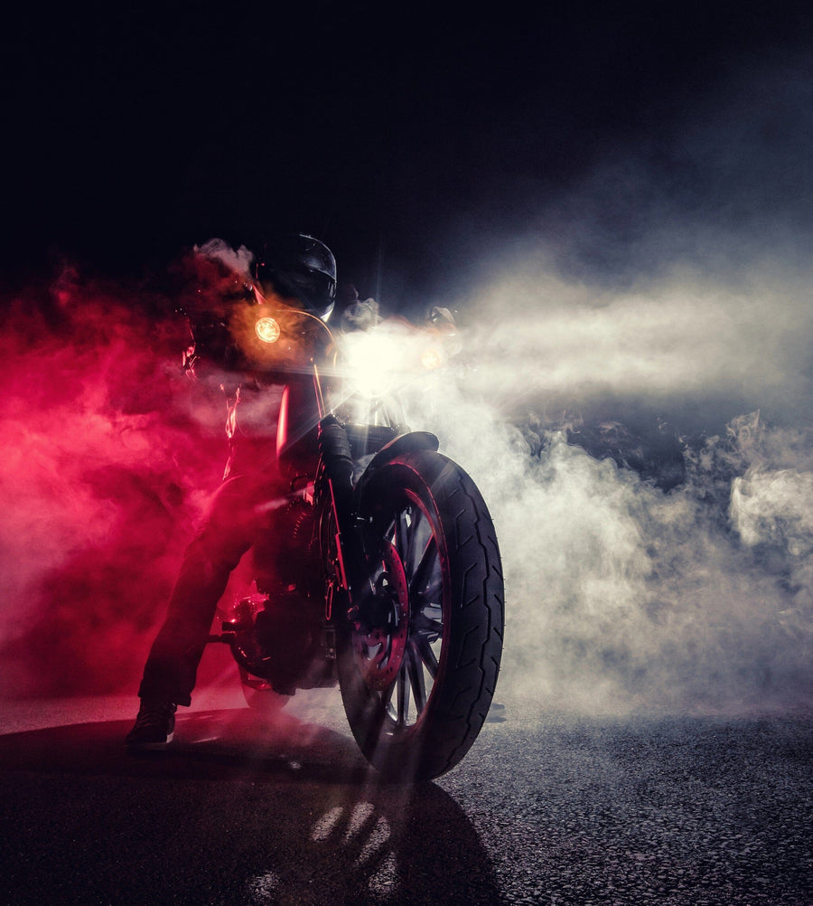 The "Motorcycle in Smoke Wall Mural" by Deposit photo features a dramatic scene of a motorcycle with bright headlights piercing through dense smoke, angled slightly. The rider's dark helmet is barely visible amidst red and white lights contrasting against the dark backdrop, while the road reflects the light.