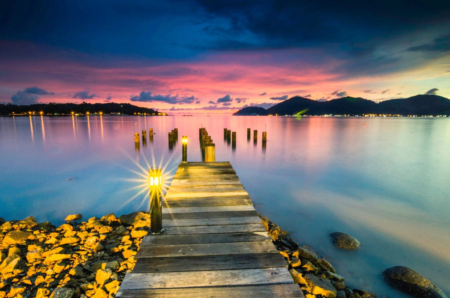 The "Old Bridge Wall Mural" by Photo features a wooden pier stretching into a serene lake at sunset, with luminous lanterns along the path. The vibrant sky in purple, pink, and orange hues reflects on the water beside silhouetted distant hills, creating an ideal tranquil mural scene.