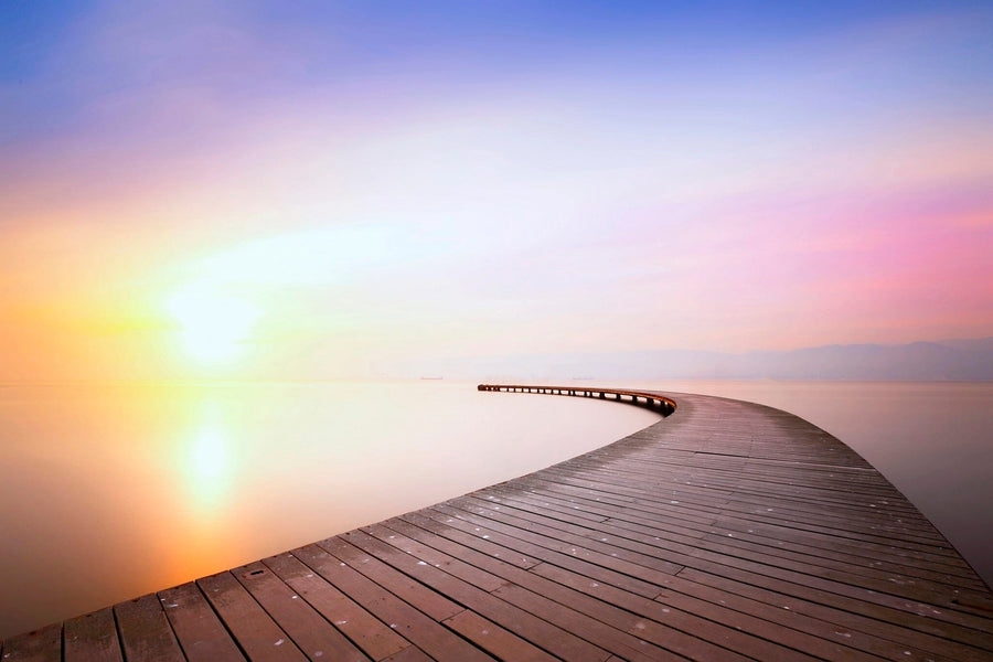 A long, curved wooden boardwalk stretches over tranquil water beneath a pastel sky at sunrise or sunset. The serene scene of our Pastel Pier Sunset Wall Mural is ideal for transforming your space with custom printed or peel and stick mural options.