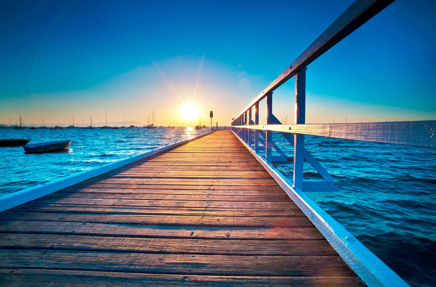 A wooden pier extends into a serene blue sea with distant boats under a clear sky. At sunset, a warm glow illuminates the scene, much like the "Path to Sea" wall mural that invites you to immerse yourself in your own peaceful sunset.