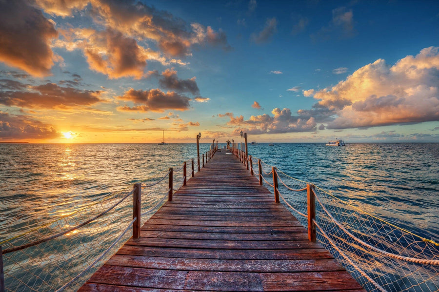 A wooden pier reaches into a calm ocean under a vibrant sunset, reflected in shades of the Peaceful Ocean View Wall Mural. The sky is an artful tapestry of orange, pink, and blue clouds, with rope fencing on the pier and a sailboat afar, capturing nature's perfect peel-and-stick mural.