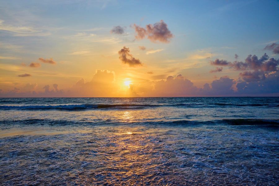 The vibrant sunset over the ocean, with its partially hidden sun casting orange and yellow hues through sparse clouds, mirrors the serene scene depicted on the Phuket Beach Wall Mural. Gentle waves reflect warm colors towards the shore, evoking a custom peel and stick mural's essence.