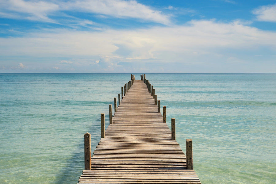 The Pier 8 Wall Mural depicts a wooden pier stretching into a serene, turquoise sea under a blue sky with scattered clouds, transitioning to deep blue farther out for a tranquil, summery feel.