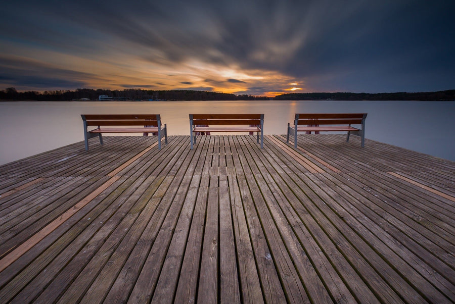 Enhance your space with the Photo "Pier and Benches Wall Mural," featuring a tranquil lakeside sunset with dramatic clouds and a colorful horizon, reflecting on smooth water. The wide wooden dock with benches offers a serene view, perfect for peel-and-stick installation.