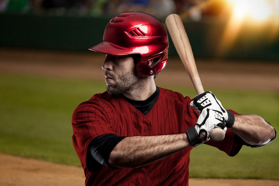 A baseball player in a shiny red helmet and pinstriped jersey stands ready to bat with a wooden bat, set against a blurred field and bright lights—perfect for decorating with the Baseball 6 Wall Mural peel and stick design.