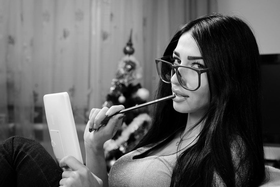 A woman with long dark hair and large glasses is seated indoors, holding a tablet and playfully biting a pen. Behind her, the blurred scene resembles the Pretty Girl in Glasses Wall Mural, with a Christmas tree adding to the ambiance. She wears a fitted top and gazes directly at the camera in this black-and-white image.