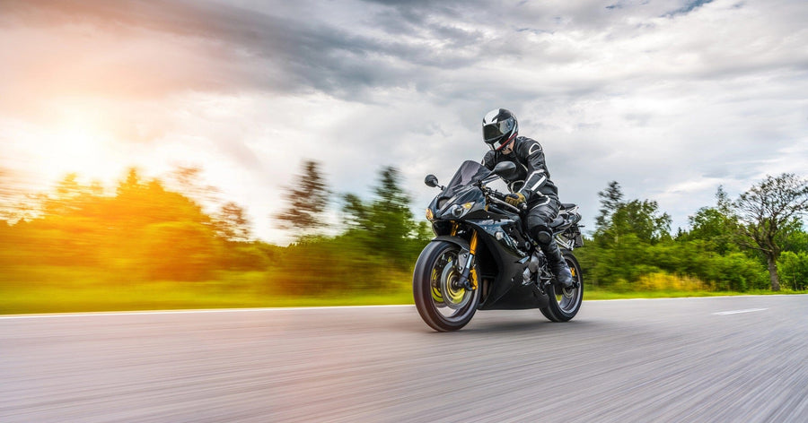 A person rides a black sport motorcycle swiftly along a paved road with blurred greenery, resembling the "Riding into Motion Wall Mural." The rider in black gear and helmet is highlighted by sunlight flares on the right, while clouds fill the sky for added drama.