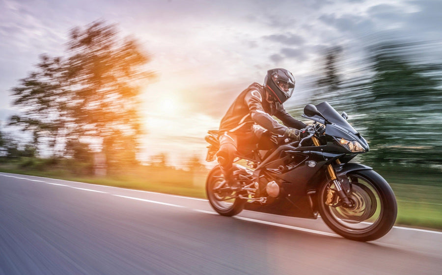 A motorcyclist in black gear zooms down a road on a sleek bike, framed by the warm glow of sunset and blurred trees, much like the Riding into the Sunset Wall Mural that brings the thrill of open roads to your walls.