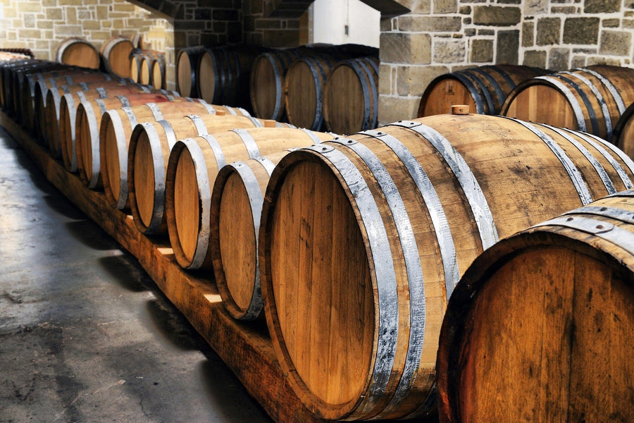 Dimly lit cellar features rows of large wooden barrels with metal bands and polished surfaces, likely for aging wine or spirits, against a stone wall. Custom printed "Wine 17 Wall Mural" by Photo adds depth and charm, while gentle light enhances the rustic atmosphere.