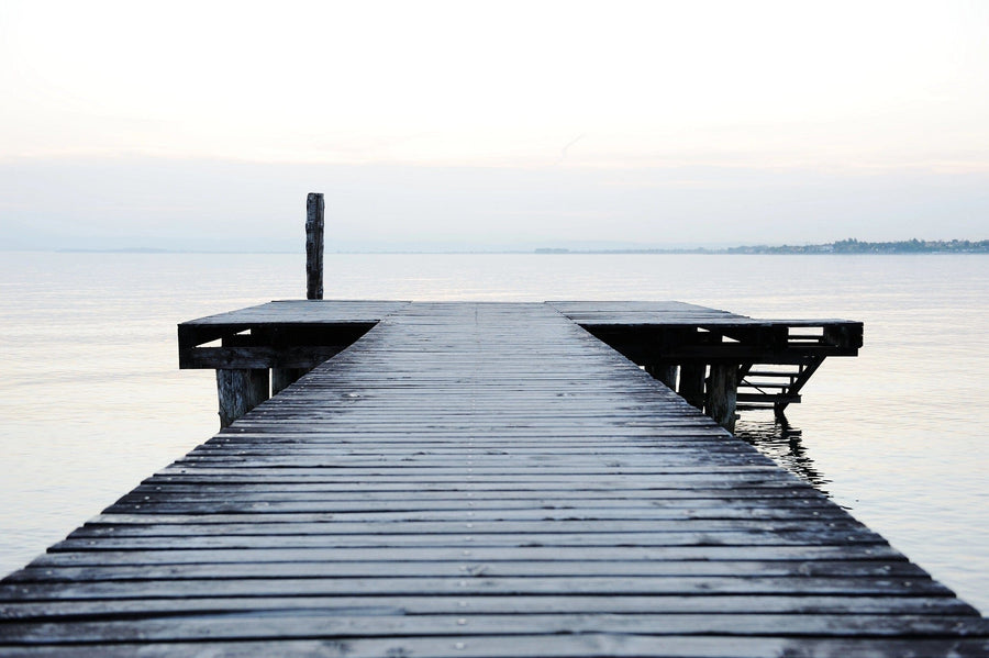 The Serene Wooden Dock Wall Mural captures a weathered pier extending into calm waters under an overcast sky, with a solitary post at its end. Faint outlines of distant land on the horizon and the sky's reflection enhance the mural's tranquil atmosphere.