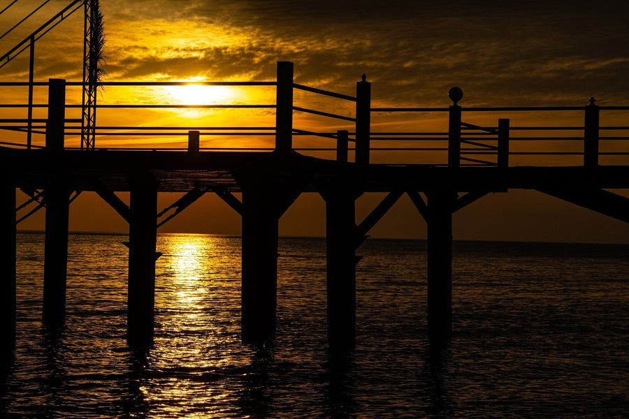 The Silhouetted Pier Glow Wall Mural features a wooden pier in silhouette over calm water at sunset, with orange and yellow skies reflected on the water—an easy peel and stick mural ideal for adding tranquil beauty to any room.