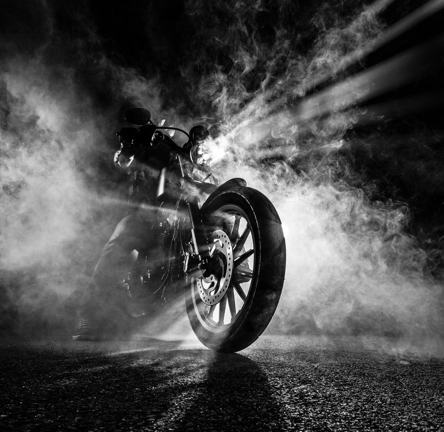 The Smoke Wall Mural by Photo features a black-and-white image of a motorcycle rider enveloped in fog. A low-angle view highlights the front wheel and dynamic spokes, while intense light beams pierce the mist, creating a dramatic and mysterious silhouette.