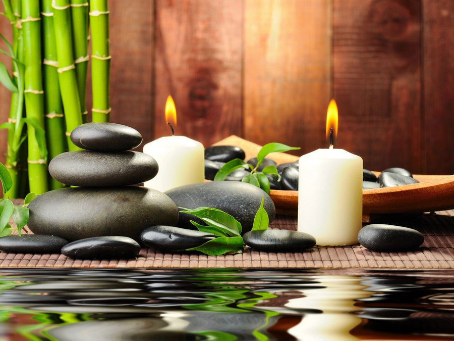 The photo Spa Wall Mural creates a tranquil spa scene with lit white candles, smooth black stones on a bamboo mat, and green bamboo stalks reflected in the water below.