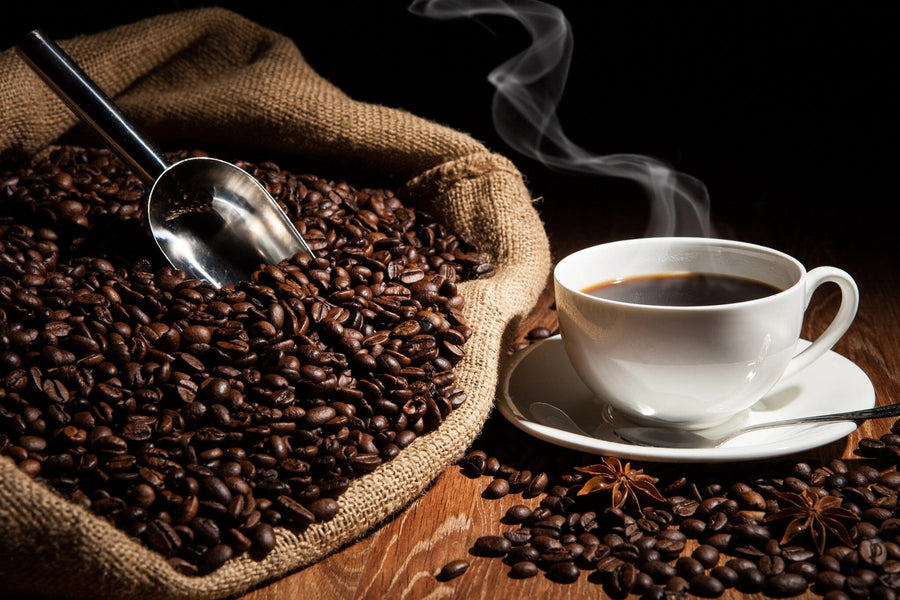 A Steaming Cup of Coffee Wall Mural by Photo features a cup beside a burlap sack brimming with roasted beans, a metal scoop partially buried, and two star anise nearby—all warmly lit to showcase the rich textures of this cozy scene.