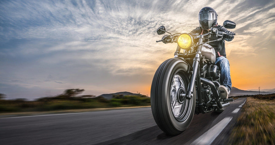 Wearing a black helmet and jacket, the motorcyclist rides a classic bike on an open road at sunset in this "Sunset Biker Wall Mural," where the headlight beams through an orange and blue sky with wispy clouds framing the scene, epitomizing speed and freedom against grassy hills.