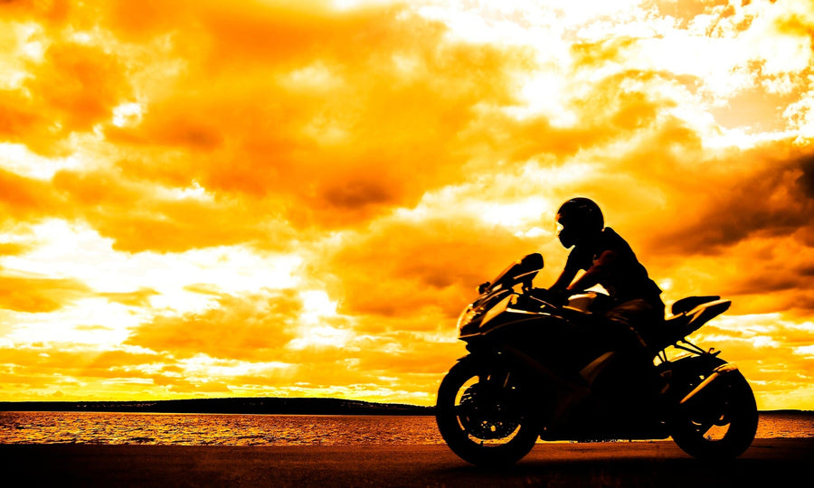 A silhouetted motorcyclist rides under a vibrant orange and yellow sunset sky, evoking the essence of the "Sunset Chase Wall Mural." Dramatic clouds and a body of water enhance the striking contrast between the dark silhouette and the bright, mural-like horizon.