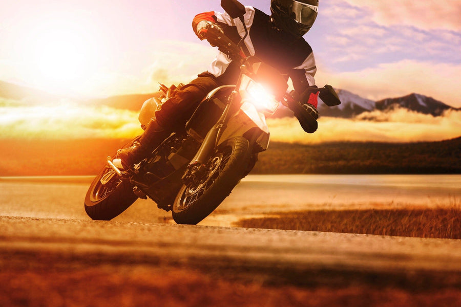 A motorcyclist in black and white gear leans sharply while cornering near a lake at sunset, creating a scene reminiscent of the Sunset Drift Wall Mural, with mountains and clouds in the distance adding to the dramatic atmosphere.