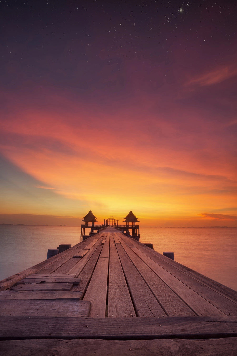 The Sunset Over Pier Wall Mural by features a wooden pier leading to a gazebo-like structure set against a vibrant sunset sky, with orange, pink, and purple hues dotted with stars, capturing nature's serene beauty reflected in the calm water below.