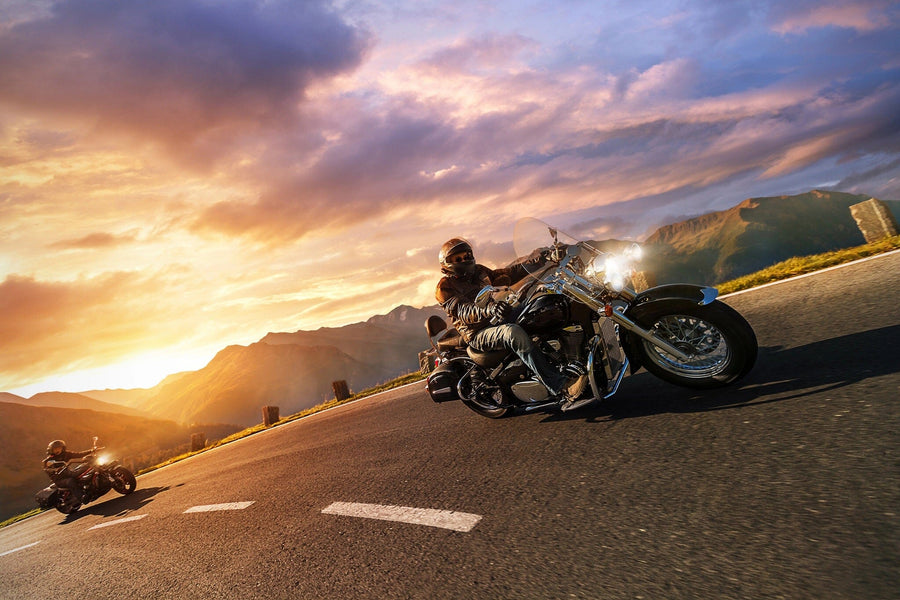 Two motorcyclists cruise along a mountain road at sunset, the lead on a large cruiser. The vibrant sky, resembling the "Sunset Ride Wall Mural" by, glows with oranges and pinks, illuminating the landscape and highlighting distant mountains like custom printed murals.