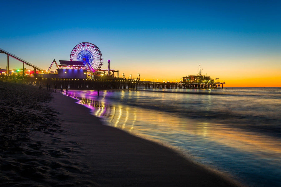 Experience the "Sunset at the Pier Wall Mural", showcasing a vibrant beach scene with a glowing Ferris wheel. The sky shifts from deep blue to orange, reflecting on water and wet sand, akin to removable wallpaper filled with colorful lights and a serene shoreline.