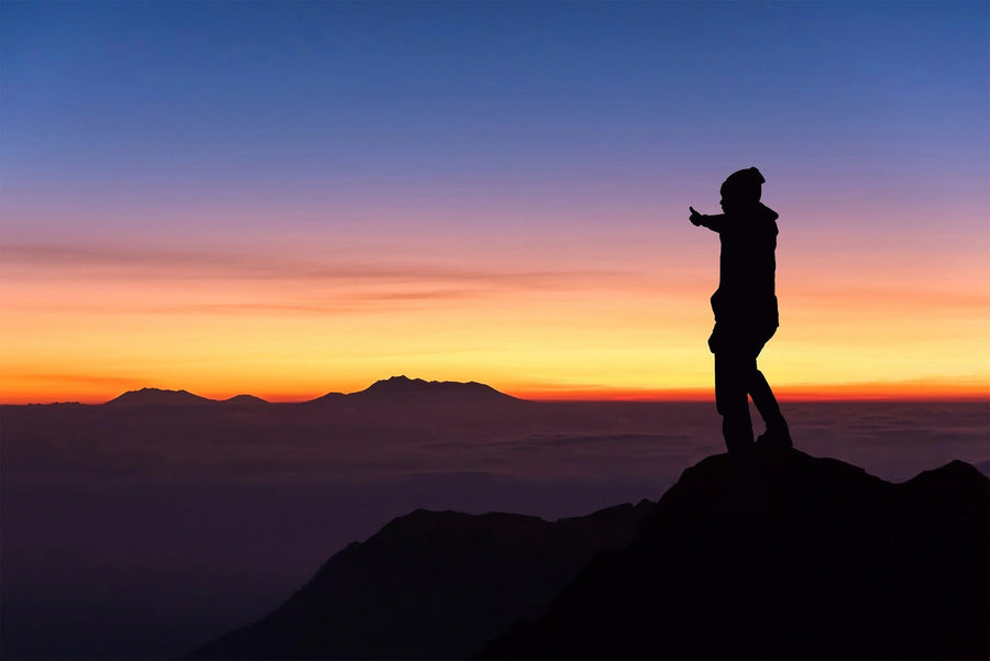 A silhouette of a person in a beanie and jacket stands on a mountain peak, pointing towards distant ranges. The scene resembles the Thumbs Up Wall Mural  with blues and oranges suggesting sunrise or sunset, capturing nature's grandeur as clouds hover below.