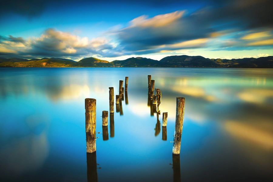 The Pier 5 Wall Mural showcases a serene lake scene with wooden posts emerging from calm, reflective turquoise waters. Dark hills meet soft sunlight under partly cloudy skies in the backdrop, while a long exposure effect blurs clouds and enhances reflections for a peaceful ambiance.