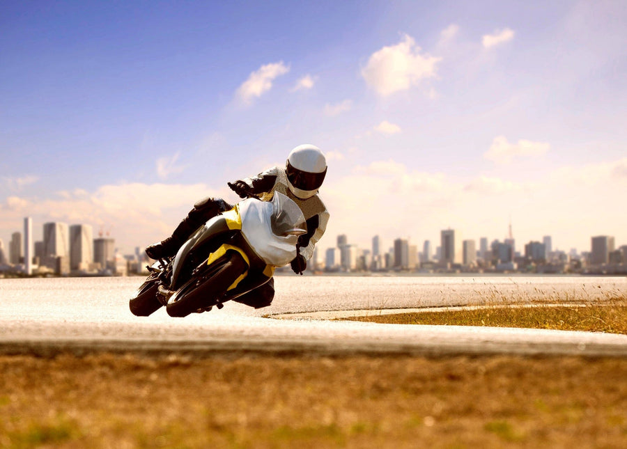 A motorcyclist in a white helmet and suit leans into a turn on a paved road. The predominantly white and yellow bike mirrors the sleek design of the Urban Speed Chase Wall Mural. A city skyline is visible under the clear blue sky, with dry grass lining the roadside in front.