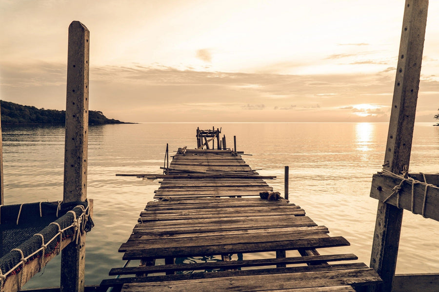 The Weathered Dock at Sunset Wall Mural features a wooden pier with missing planks stretching over calm water at sunset, bringing a tranquil golden sky scene to your space as a custom printed, removable wallpaper.