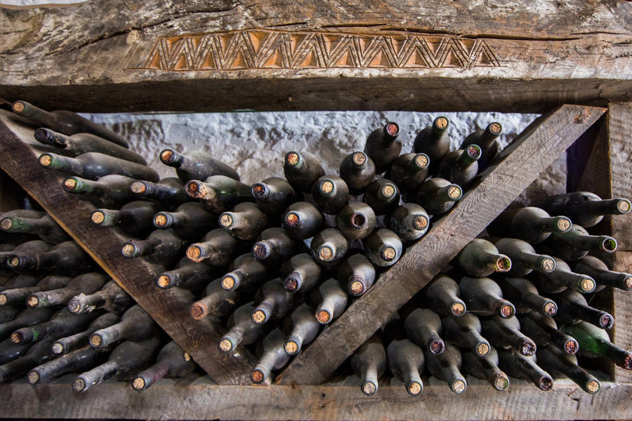 A rustic wine rack holds dusty wine bottles arranged in a crisscross wooden structure with necks facing outward. Above the rack, a decorative wooden panel complements Deposit photo's elegant "Wine Bottles Covered in Dust Wall Mural" adorning the stone wall.