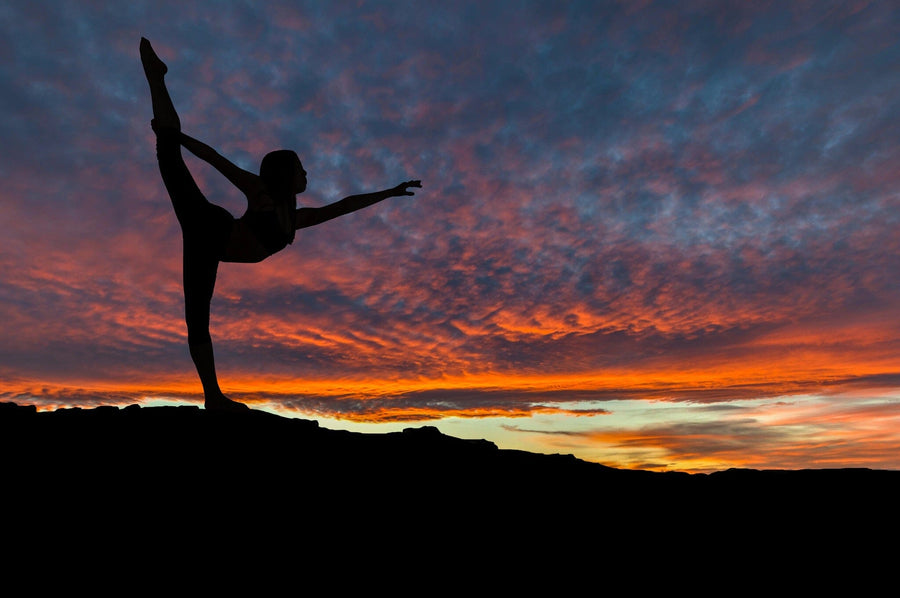 A woman performs a balancing yoga pose on one leg at sunset, her silhouette set against a vibrant backdrop of orange, pink, and purple. This dramatic view mirrors the serene energy captured in Deposit Photo's Woman Stretching Wall Mural.