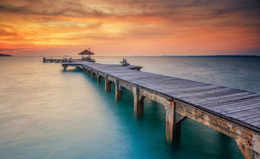 A tranquil Wood Bridge Wall Mural from Deposit Photo features a pier stretching over calm turquoise waters toward an open shelter, set against a sunset sky of orange, pink, and purple hues, casting a warm glow.