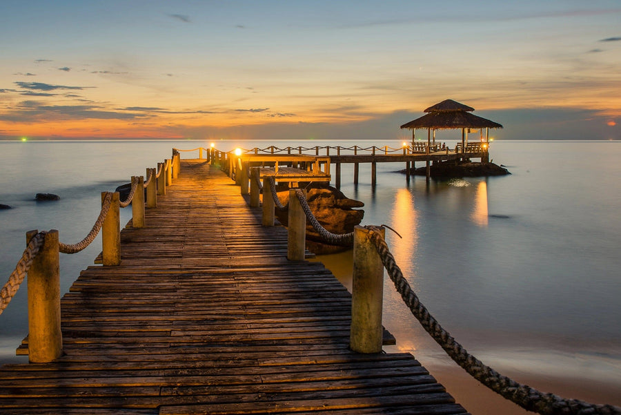 A tranquil scene depicted in the Wooden Bridge Landscape Wall Mural by features a wooden pier over calm water leading to a thatched-roof gazebo. The sunset sky's orange and blue hues, along with soft light on the pier, evoke serenity, while rocks enhance the natural beauty.