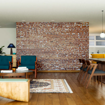 A modern living and dining area features a Mismatched Bricks Wall Mural, blue armchairs, green sofa, round coffee table, wooden dining table for six, pendant lights, rugs, hardwood floors, and a kitchen with white cabinets in the background.