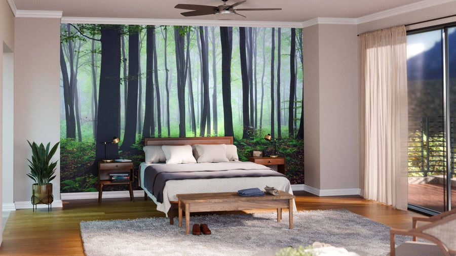 A cozy bedroom showcases a bed with white and gray bedding, wooden furniture, a gray rug, and the Misty Autumn Forest Wall Mural. Sunlight filters through glass doors with sheer curtains, while a potted plant accents the corner.