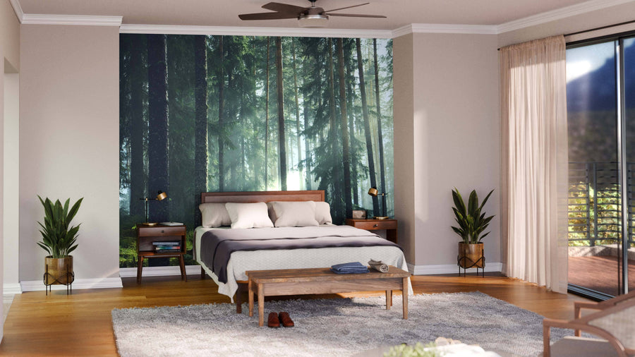 A modern bedroom highlights the Misty Forest Stand Wall Mural behind a wooden bed. Two bedside tables with lamps and plants sit on wood floors, a bench rests on a light rug, and sunlight streams through sheer curtains beneath a ceiling fan.