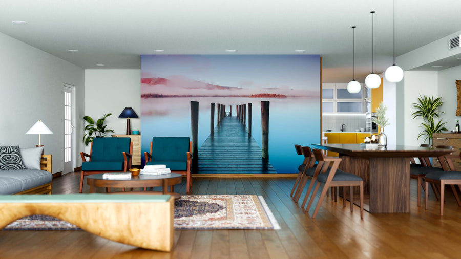 Modern open living-dining room with wood floors, blue chairs, the "Misty Lake Morning Wall Mural" depicting a dock over calm water, pendant lights, rugs, plants, dining table for six, and a kitchen with yellow accents in the background.