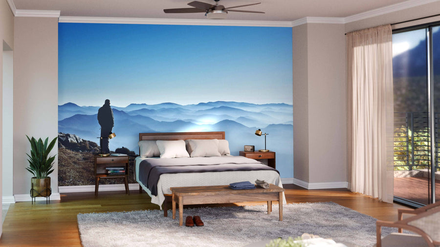 A modern bedroom features wood furniture, light walls, and the Misty Mountain Hills Wall Mural showing a person on a peak with blue misty hills. Sunlight shines through glass doors, highlighting the rug, bench, and indoor plant on hardwood floors.