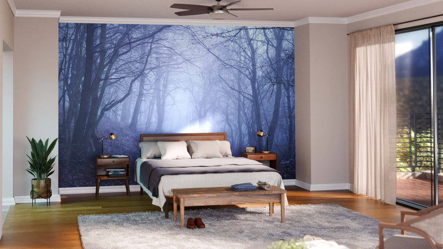 A modern bedroom with wood furniture and a gray bedspread features the Misty Spooky Purple Forest Wall Mural. Large windows with sheer curtains let in natural light, while a plant, rug, and bench complete the serene space.