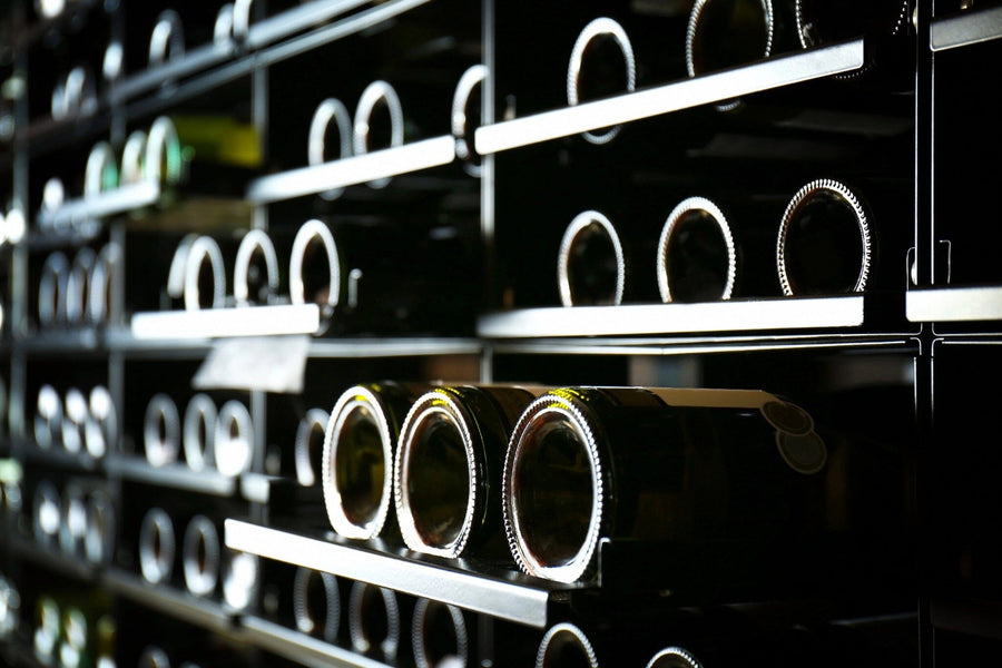 Modern café interior with wooden tables, potted plants, and large windows. A Modern Wine Rack Wall Mural of stacked wine bottles adds character, while pendant lights hang from the ceiling and wine shelves line the right wall.