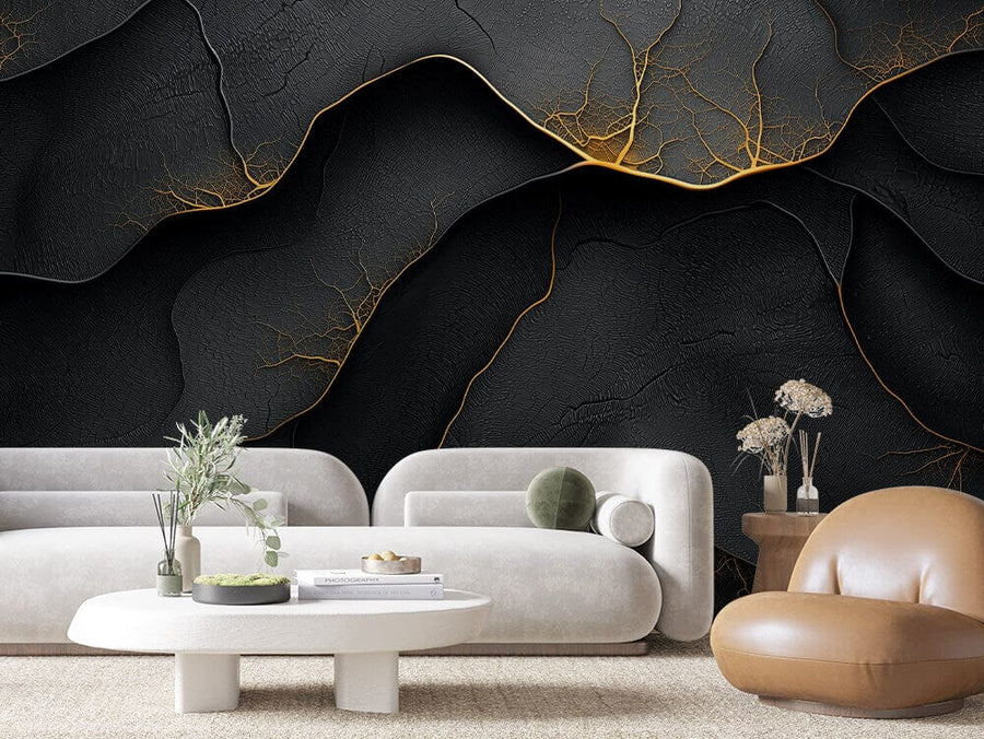 A modern living room showcases the Molten Fractures Wall Mural, with cracked black textures and gold accents akin to abstract veins. It complements two white sofas, a brown leather chair, an oval coffee table with books and plants, and a round side table with dried flowers on a beige rug.