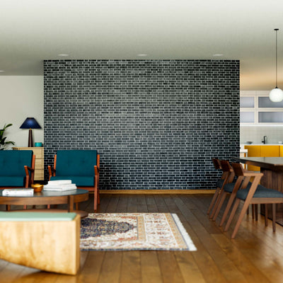 A modern living and dining area features teal armchairs, a gray sofa, a wooden coffee table, and a dark accent wall highlighted by the Monochrome Brick Pattern Wall Mural, with houseplants and an open kitchen with yellow cabinets in the background.