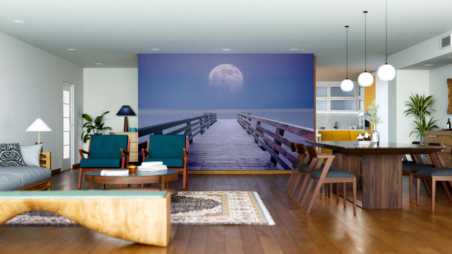 A modern living and dining area with wooden floors, contemporary furniture, plants, soft lighting, and calming blue, green, and wood tones is highlighted by the Moonlit Horizon Wall Mural featuring a moonlit pier over water.