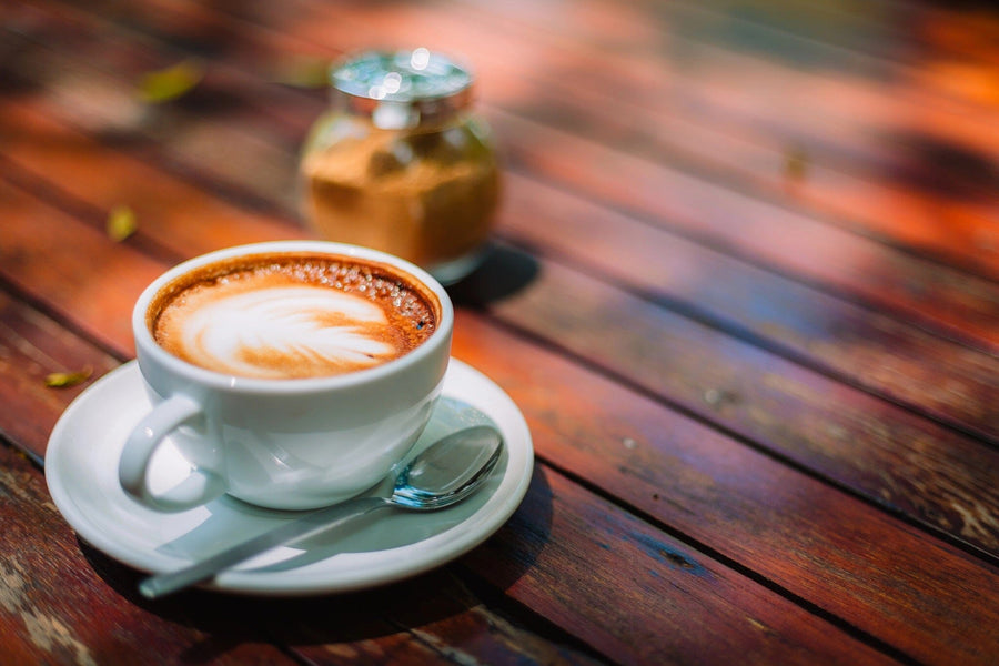 A modern café with wooden tables, black stools, and hanging lights showcases the Morning Coffee Wall Mural—a large image of a cappuccino in a white cup on wood. Small plants complete the inviting, charming atmosphere.